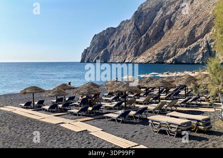 Touristen entspannen sich am schwarzen Sandstrand von Perissa, Santorin, Griechenland, und genießen die Sonne und das Meer unter Strohschirmen mit dem imposanten Mesa Vouno Moun Stockfoto