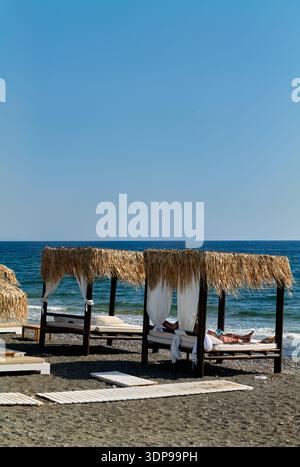 Ein Urlauber genießt das Sonnenbaden in einer privaten strohgedeckten Cabana an einem dunklen Sandstrand mit dem ruhigen blauen Meer im Hintergrund. Stockfoto