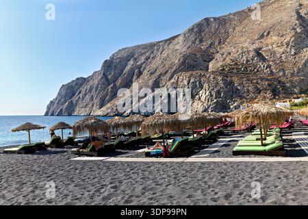 Die Menschen entspannen auf Sonnenliegen unter Strohschirmen an einem schwarzen Sandstrand in Santorin, Griechenland, und genießen den klaren blauen Himmel und die ruhige Ägäis. Stockfoto