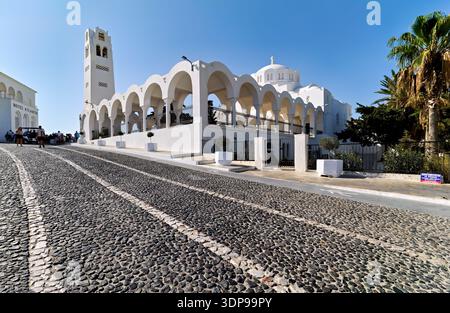 Touristen laufen auf einer Kopfsteinpflasterstraße vorbei an der weißen Metropolitan Cathedral of Thira und dem Hotel Atlantis in Fira, Santorin, Griechenland. Stockfoto