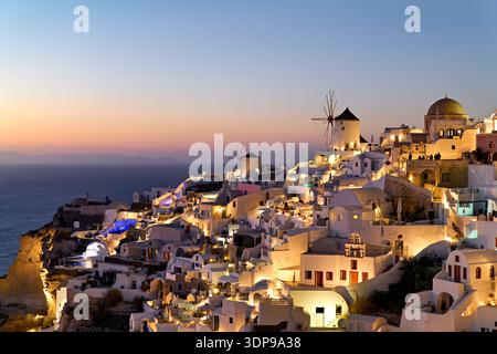 Oia, Santorini, Griechenland in der Abenddämmerung. Touristen genießen das beleuchtete, weiß getünchte Dorf und die berühmten Windmühlen mit Blick auf die Ägäis bei Sonnenuntergang. Stockfoto