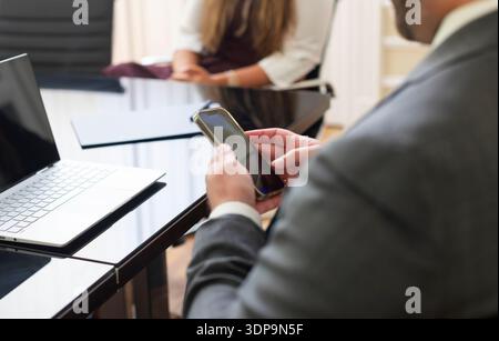 Eine Person im Anzug schaut auf ein Mobiltelefon, während sie an einem schwarzen Tisch sitzt. Eine andere Person sitzt im Hintergrund. Ein Laptop befindet sich auf dem Stockfoto