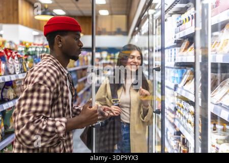 Junge, vielfältige Paare, die in einem Supermarkt in Kühlregalen stöbern, lächeln und während einer wöchentlichen Einkaufstour frische Produkte auswählen Stockfoto