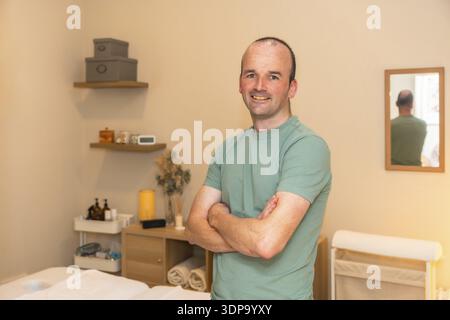 Professioneller Mann Massagetherapeut lächelnd selbstbewusst, stehend mit gekreuzten Armen in einem Therapieraum, bereit, Wellness und Gesundheitsdienstleistungen zu bieten Stockfoto