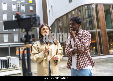 Journalistin mit Mikrofon und Gesten, während sie einen jungen Mann auf einer Stadtstraße interviewt und aktuelle Ereignisse für eine Live-Nachrichtenagentur dokumentiert Stockfoto