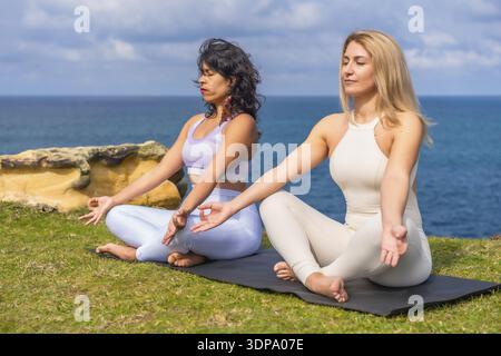 Zwei verschiedene Frauen, die in Lotus sitzen, posieren auf Yogamatten am Meer und finden Ruhe und Gleichgewicht durch achtsame Meditation für Wellness und Spirituu Stockfoto