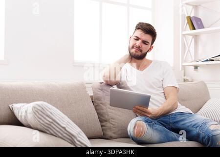 Mann, der mit Tablet auf der Couch sitzt und Nackenbeschwerden und Dehnen erlebt Stockfoto