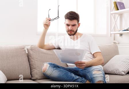 Junger Mann liest Dokumente zu Hause, während er eine Brille hält und auf einer Couch sitzt Stockfoto