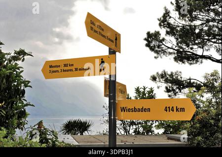 Gelbe Schilder zeigen Entfernungen zu verschiedenen Städten, Bäumen und Himmel im Hintergrund, Genfersee, Montreux, Kanton Waadt, Schweiz Stockfoto