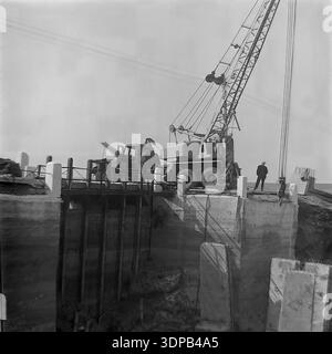 Bezirk Slawjansk, Region Donezk, Ukraine, UdSSR. Ende der 1960er - Anfang der 1970er Jahre Bau einer Betonschleuse im Betrieb Donrybkombinat. Ein schwerer sowjetischer LKW-Kran arbeitet auf einer massiven hydraulischen Struktur. Ein Arbeiter überwacht den Installationsvorgang vom Rand des Damms aus. Blick auf die tiefe Fundamentgrube und die Stahlbetonwände während der Reparaturarbeiten. Ausbau der Infrastruktur für die Fischzucht. Eine Industrieszene der Ingenieure und Arbeit im friedlichen Donbass vor dem Krieg. Stockfoto