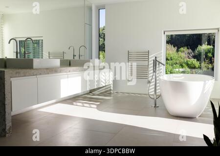 Freistehende, ovale, weiße Badewanne und schwebender Kosmetikbereich glänzen unter Sonnenlicht und bieten Blick auf den Garten Stockfoto