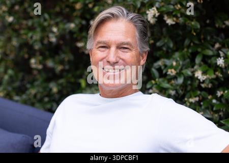Reifer Mann sitzt in dunkelblauer Lounge mit marineblauem Kissen im Garten und trägt ein weißes T-Shirt und lächelt Stockfoto