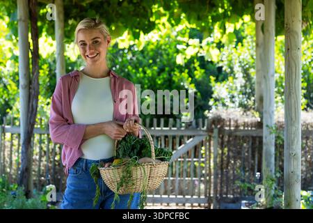Frau, die unter Pergola im Garten steht und Korbkorb mit Produkten hält, mit rosa Hemd Stockfoto