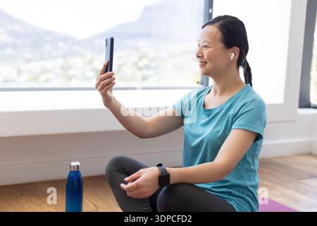 Asiatische Frau sitzt auf lila Matte zu Hause am Fenster, oben, hält Telefon, Ohrstöpsel, Kopierraum Stockfoto