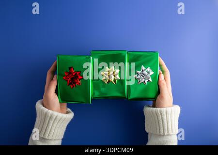 Foto von der ersten Person von oben mit weiblichen Händen, die drei glänzend grüne Geschenkboxen mit Schleife auf isoliertem blauem Hintergrund halten Stockfoto