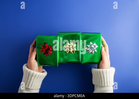 Foto von der ersten Person von oben mit weiblichen Händen, die drei glänzend grüne Geschenkboxen mit Schleife auf isoliertem blauem Hintergrund halten Stockfoto