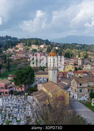 Senkrechter Blick aus der Luft auf das Dorf Potamos in Korfu Griechenland mit dem historischen Kirchturm und dem Friedhof mit weißen Marmorgräbern unter einem bewölkten s Stockfoto