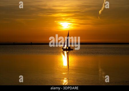 Ein kleines Segelboot mit goldenem Sonnenuntergang, der sich auf dem ruhigen Wasser des West Kirby Marine Lake spiegelt Stockfoto