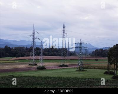 Hochspannungs-Übertragungstürme und Stromleitungen werden durch eine ländliche Landschaft unter einem bewölkten Himmel mit weit entfernten Bergen gesehen. Stockfoto