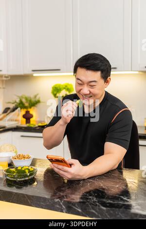 Asiatischer Mann in der Küche isst Brokkoli-Snack und überprüft Gesundheitsnachrichten auf orange Smartphone. Ein erwachsener Mann mittleren Alters genießt kalorienarme Snacks drinnen und fühlt sich motiviert, den Ernährungsplan und die Änderung des Lebensstils zu befolgen. Stockfoto