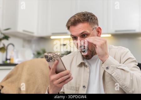 Ein junger Mann zu Hause liest mit einer Brille winzige Texte auf dem Smartphone und staunt über die Geschichte und Neuigkeiten. Der kaukasische Kerl auf dem Sofa sieht starke Aufmerksamkeit im Boten, zeigt Freude Erleichterung und helle Wow-Überraschung auf einmal Stockfoto
