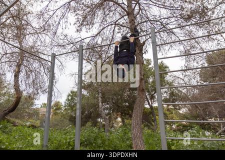Junger Mann, der einen umgekehrten Hang an einer horizontalen Bar während eines Calisthenics-Workouts im Freien in einem von Kiefern umgebenen Waldpark durchführt Stockfoto