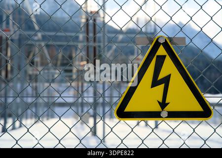 Dreieckiges gelbes Warnschild für Hochspannung am Zaun, Warnung vor Stromschlag, hinter der Transformatorstation unscharf, elektrisches Sicherheitssystem Stockfoto