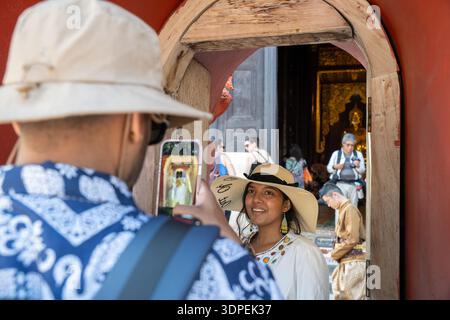 Ein Schuss über die Schulter eines Mannes mit Hut, der ein Smartphone-Foto einer lächelnden Frau macht. Sie sind Touristen, die einen lebendigen rot-goldenen Tempel i besuchen Stockfoto