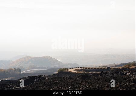Eine gewundene Bergstraße führt durch dunkles vulkanisches Gelände auf dem Ätna, umgeben von schwarz-weißen Barrieren, die den scharfen Kurven folgen. Gezackt l Stockfoto