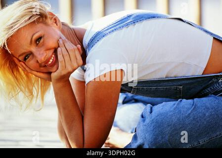 Porträt einer attraktiven blonden Frau sitzt gern auf dem Boden mit den Händen auf den Wangen und lächelt in die Kamera. Sie trägt blaue Overalls und ein weißes T-Shirt Stockfoto