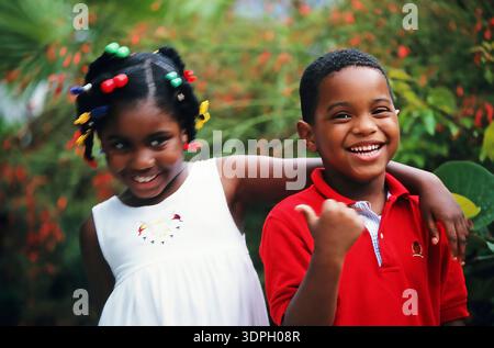 Bruder und Schwester stehen dicht beieinander in einem Garten. Ein Mädchen trägt ein weißes Kleid und schaut seitlich, während ein Junge in Rot lächelt und die Daumen hoch zeigt. Br Garten Stockfoto