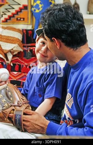 Ein Latino Vater und Sohn haben Spaß zusammen, während sie einen Baseball im Wohnzimmer anschauen. Der Vater lächelt den Sohn an, der einen Handschuh hält Stockfoto