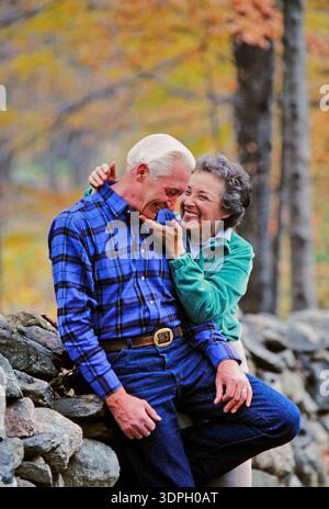 Ein älteres Paar sitzt auf einer Steinmauer mit Bäumen im Hintergrund. Sie lachen und genießen die Gesellschaft des anderen in einer hellen Herbstszene Stockfoto