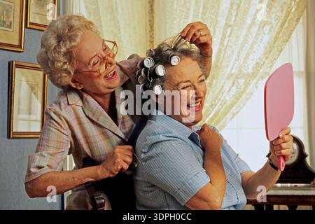 Zwei ältere Frauen teilen sich einen freudigen Moment in einem hellen Raum. Eine Frau frisiert die Haare der anderen mit Rollen, während sie lächelt und ihr Spiegelbild bewundert Stockfoto
