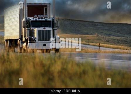 Große Lkw fahren auf offener Straße, umgeben von Gras und Hügeln, während die goldene Stunde hell wird Stockfoto