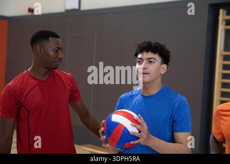 Verschiedene männliche Teamkollegen trainieren Teenager, die im Fitnessstudio Tricolor Volleyball halten und rot-blaue Hemden tragen Stockfoto