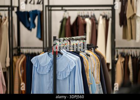 Farbenfrohe Damenbekleidung auf schwarzen Metallregalen in einem Boutique-Geschäft. Blaue Blusen im Fokus mit verschiedenen Kleidungsstücken, die im Hintergrund hängen, schaffen DE Stockfoto
