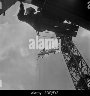 Ein außergewöhnliches Archivfoto aus den späten 1960er Jahren aus einer extrem niedrigen Perspektive auf der Baustelle des Slawjanskaja Thermal Power Plant in Nikolaevka, Donbass. Der Schuss blickt direkt nach oben und fängt einen gewagten Höhenarbeiter ein, der am Himmel hängt und Montageaufgaben an der monumentalen Struktur durchführt. Neben ihm reicht das komplizierte Gitter eines riesigen Turmdrehkrans senkrecht in die Wolken. Dieses historische Bild dokumentiert perfekt den Mut und das technische Können der friedlichen Industrialisierung. Stockfoto
