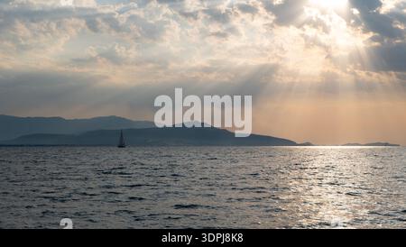 Segelboot auf dem ruhigen Meer mit Sonnenstrahlen und Bergen bei Sonnenuntergang Stockfoto