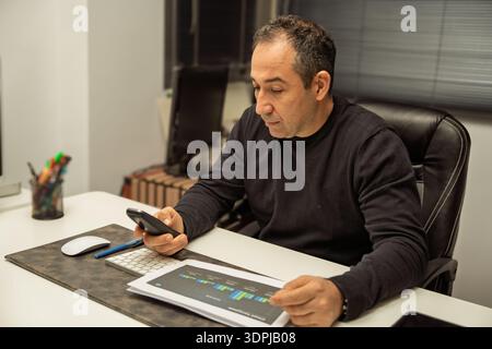 Mann, der Finanzpläne auf Dokumenten und Smartphone überprüft, während er in seinem Büro arbeitet Stockfoto