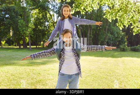 Glücklicher Vater, der ein lächelndes Kind auf den Schultern im sonnigen Park trägt, während er Flugzeug spielt und Spaß hat Stockfoto