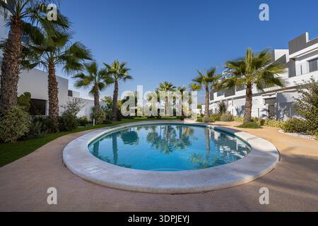 Moderne weiße Apartmentgebäude, die einen runden Gemeinschaftspool mit Palmen und landschaftlich gestaltetem Grün unter klarem blauem Himmel umgeben. Stockfoto