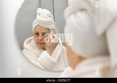 Frau mittleren Alters mit Handtuch auf dem Kopf, die auf den Badezimmerspiegel blickt, Selbstpflege und Entspannung. Kopierbereich Stockfoto