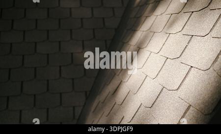 Nahaufnahme der Asphaltdachschindeln, die ein strukturiertes Muster erzeugen. Sonnenlicht beleuchtet einen Teil der dunkelgrauen Oberfläche Stockfoto