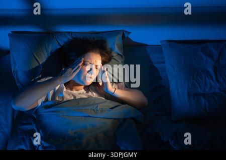 Frau mittleren Alters im Bett mit Migräne und Schlaflosigkeit in der Nacht, weibliche Kopfschmerzen und Schlafstörungen im Schlafzimmer mit Mondlicht Stockfoto