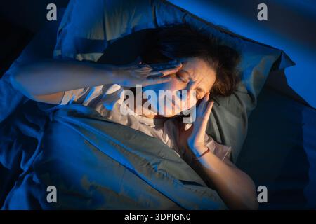 Frau mittleren Alters im Bett mit Migräne und Schlaflosigkeit in der Nacht, weibliche Kopfschmerzen und Schlafstörungen im Schlafzimmer mit Mondlicht. Stockfoto