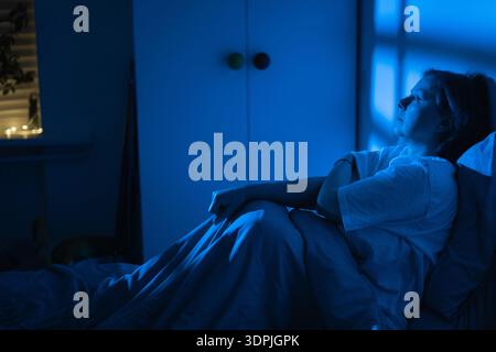 Erschöpfte Frau mittleren Alters, die im Bett sitzt und nachts durch das Fenster schaut, Frau, die an Schlaflosigkeit und Arbeitsstress leidet, fühlt sich einsam und einsam Stockfoto
