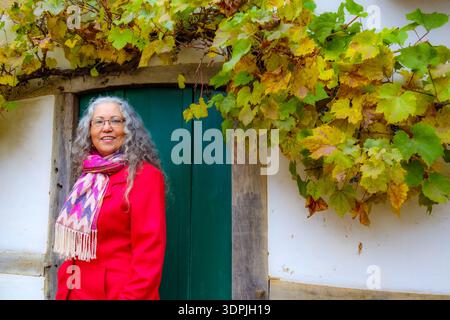Taille-up Porträt einer eleganten lächelnden Reifen Frau, die vor grüner Holztür steht, graue Haare, Brille, rosa Mantel und Schal, Kletterpflanze auf t Stockfoto