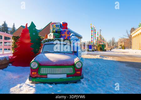 Roter Oldtimer mit bunten Weihnachtsgeschenkboxen auf dem Dach und einem freundlichen Schneemann in einem sonnigen Park. Festliche Feiertagsdekoration in einer verschneiten Stadt Stockfoto