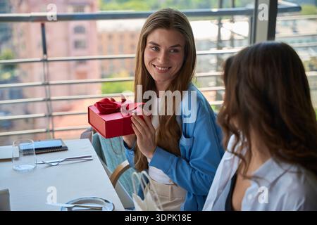 Lächelnde junge Frau feiert, hält rote Geschenkbox im Restaurant mit ihrem Freund Stockfoto